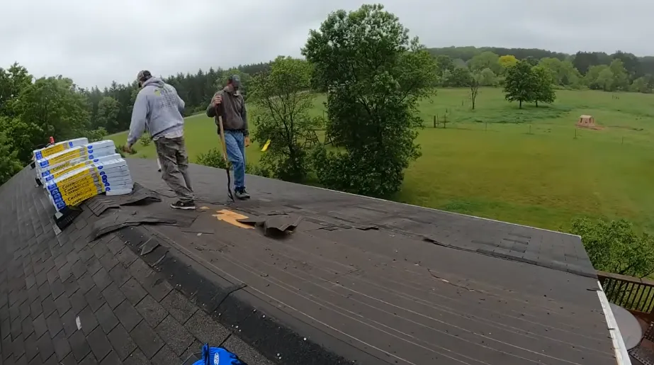 Two roofers starting a shingle tear-off beside staged shingle bundles